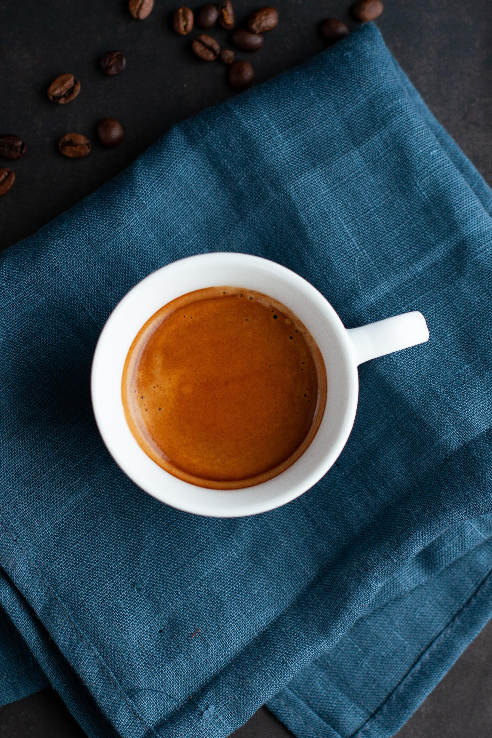 white coffee cup holding espresso on top of a blue cloth napkin surrounded by espresso beans
